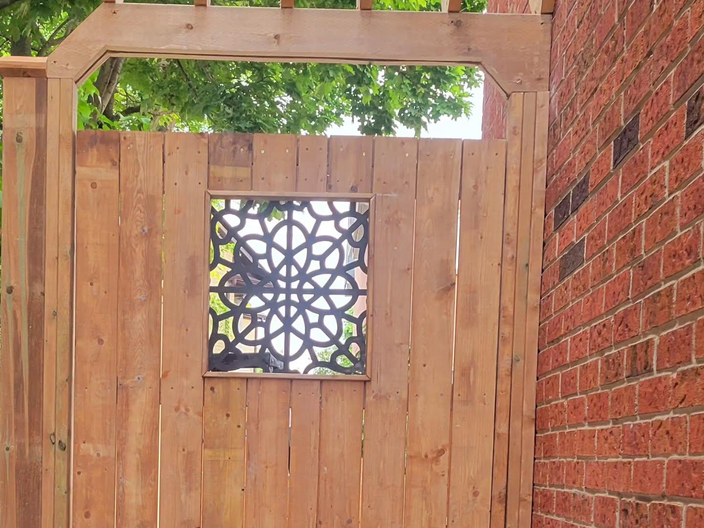 Newly installed wooden privacy fence gate with decorative black metal scrollwork insert, positioned next to a red brick residential home, with green tree foliage visible above the gate.