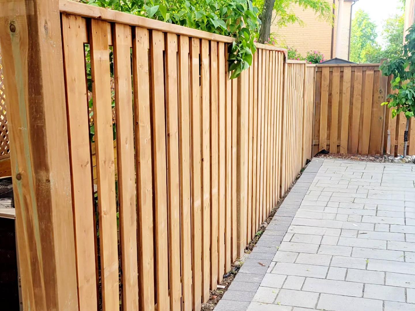 Newly installed light wood vertical slat privacy fence bordering a residential paved patio, with green foliage and a house in the background