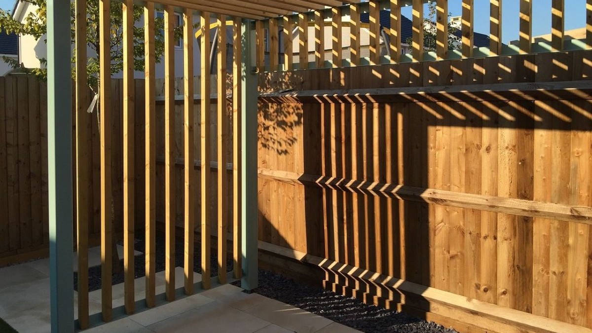 Sunlit residential backyard featuring a modern wooden slatted pergola structure, warm-toned wooden privacy fence, gravel landscaping border, and paved patio, with dappled shadows cast across the fence by the slatted design.