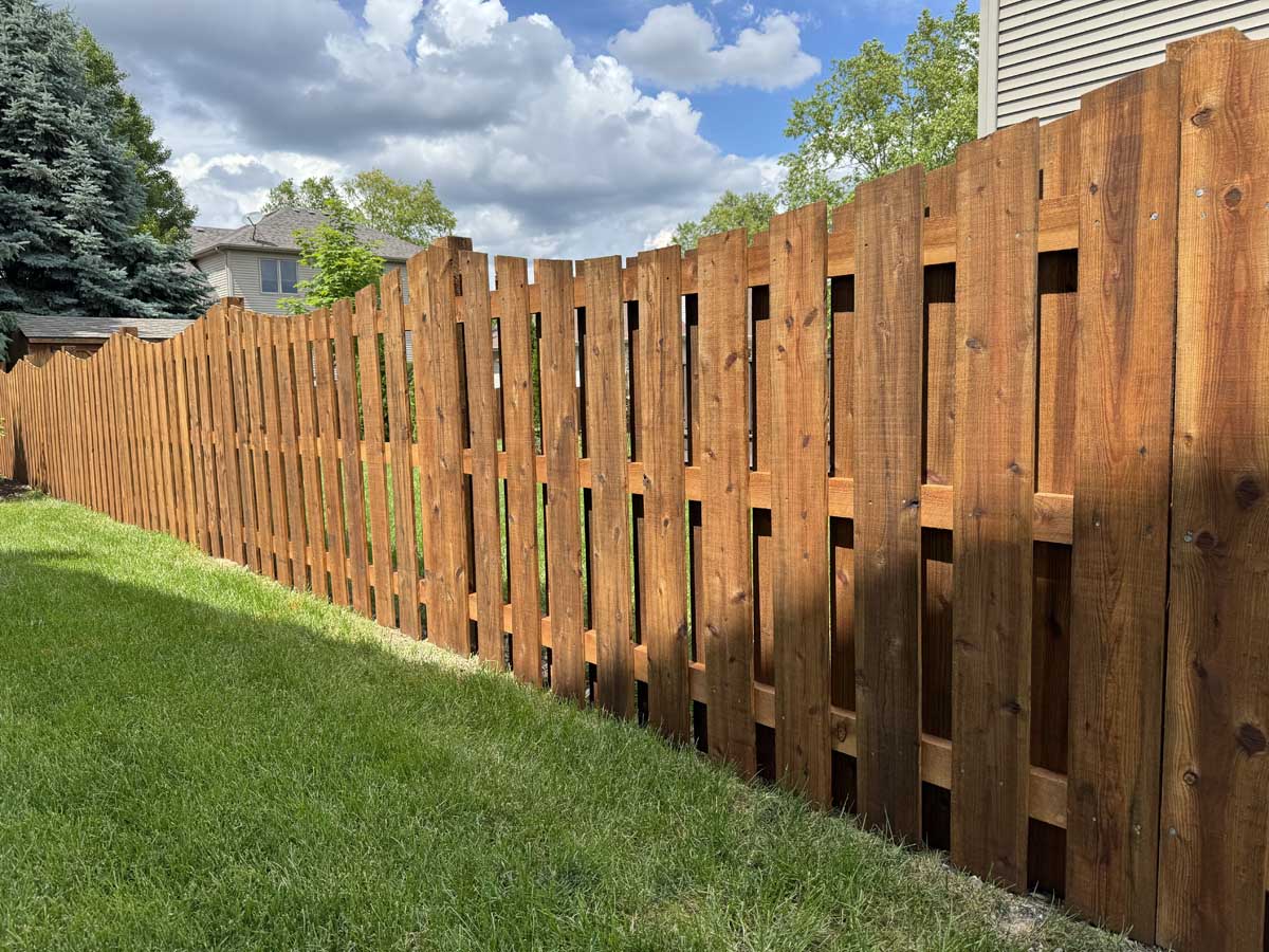 Newly installed stained wooden privacy fence with a curved top edge, running along a lush green residential lawn, with suburban houses and trees visible in the background under a bright partly cloudy sky.