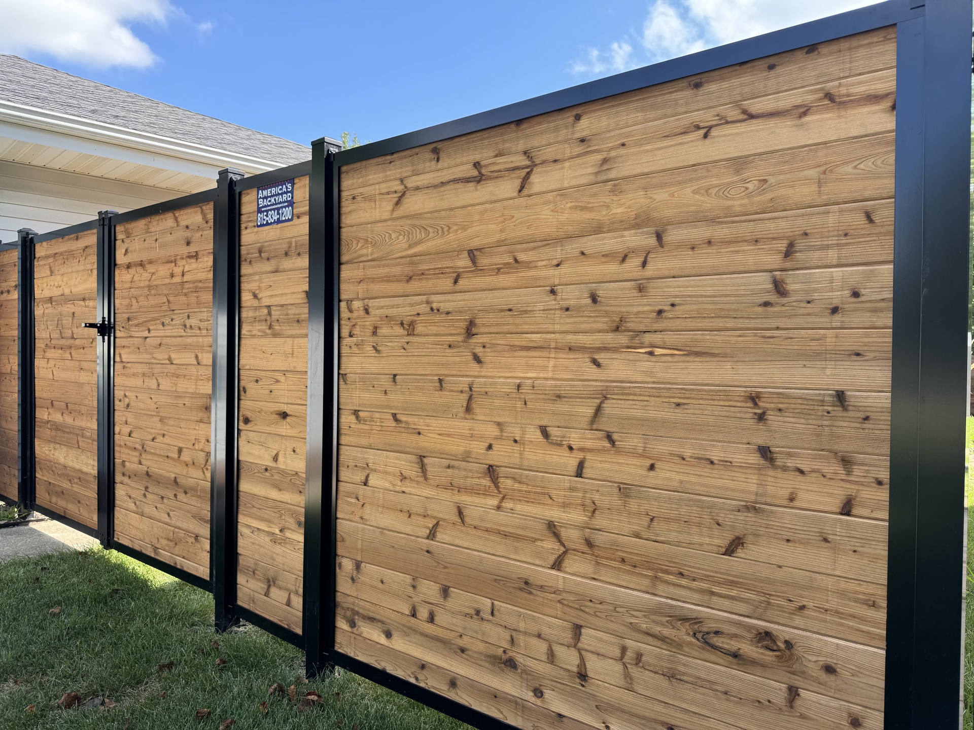 Modern horizontal wood privacy fence with black metal framing, installed in a residential yard, with a branded sign for America's Backyard fence company on one panel, under a bright sunny sky