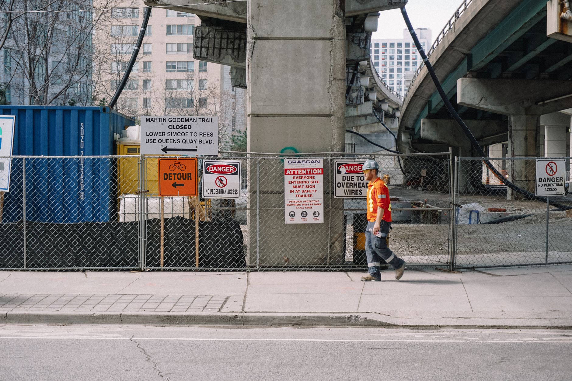 Construction worker in high-visibility safety gear walking past a chain-link construction fence at an urban road construction site under an elevated highway overpass, with detour and safety warning signs posted on the fence.